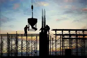 Silhueta de trabalhadores da construção civil em um andaime ao pôr do sol, com um guindaste transportando concreto, ilustrando a eficiência operacional que o BPO para construtoras pode oferecer.