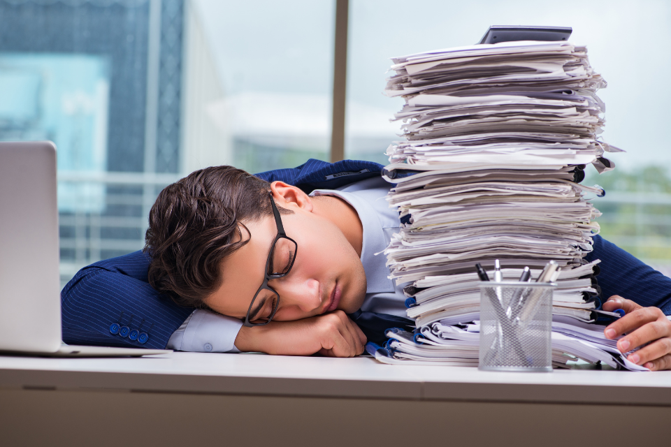 Homem exausto dormindo sobre a mesa com pilha de papéis, simbolizando excesso de trabalho manual e a importância da digitação e digitalização de documentos para reduzir tarefas repetitivas.
